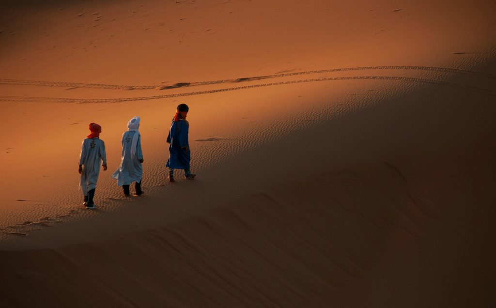 Three individuals in traditional attire walking on sand dunes during sunset.